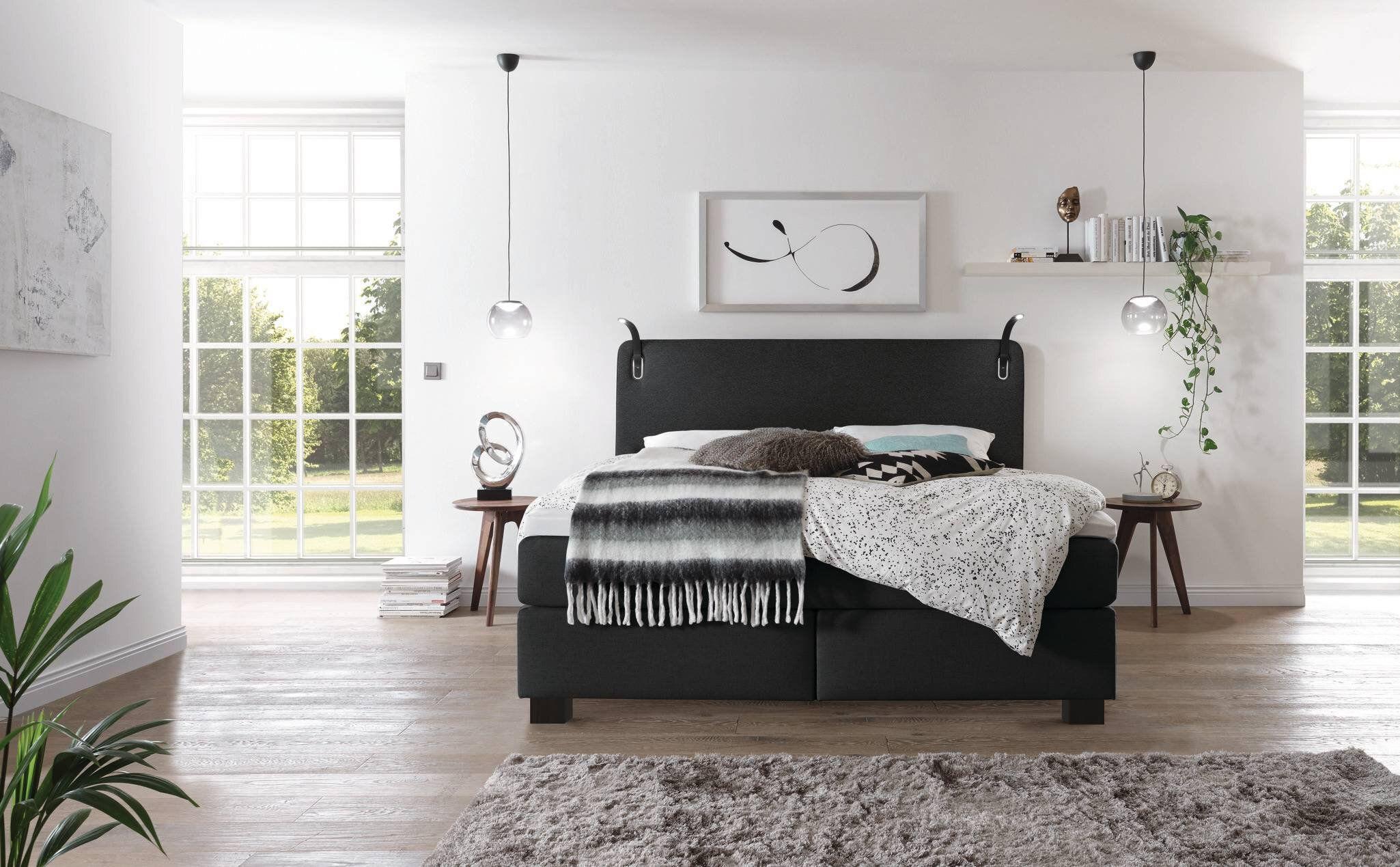 Modernes Schlafzimmer mit einem schwarzen Boxspringbett, minimalistischer Dekoration und großen Fenstern, die viel Tageslicht hereinlassen.