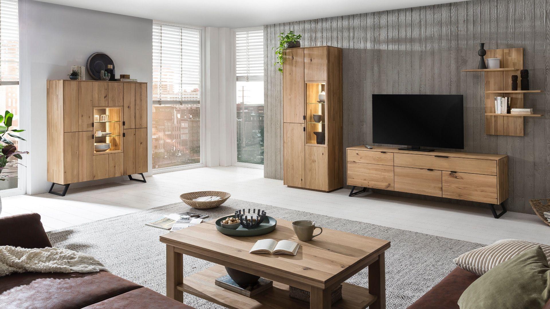 Modernes Wohnzimmer mit hellen Holzmöbeln, darunter eine Vitrine, ein Sideboard und ein Couchtisch. Ein großer Fernseher steht auf dem Sideboard, während die Einrichtung mit grünen Pflanzen und Büchern dekoriert ist.