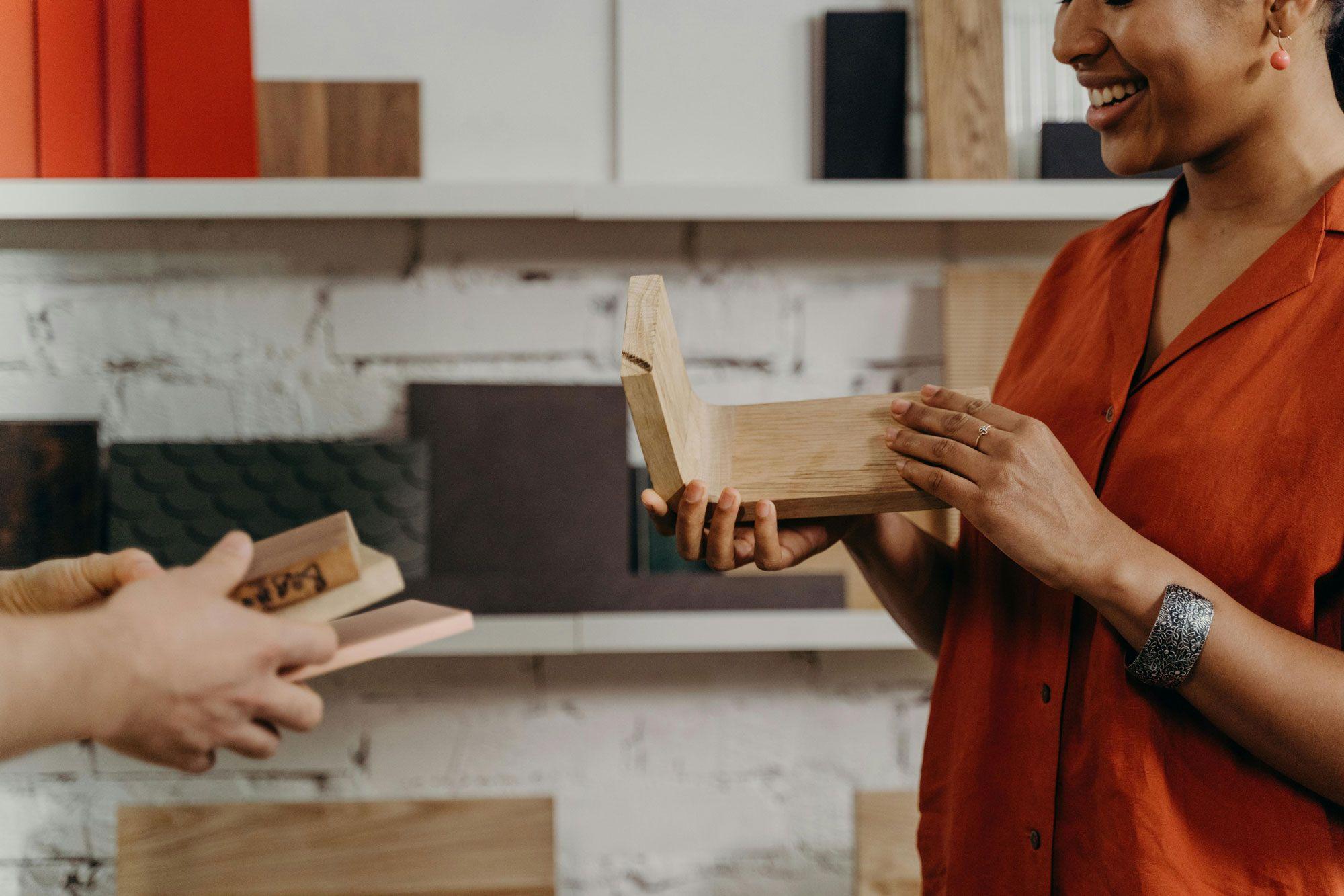 Eine Frau hält ein Holzstück in der Hand, während sie mit einer anderen Person über verschiedene Holzproben spricht, die auf einem Regal im Hintergrund angeordnet sind.