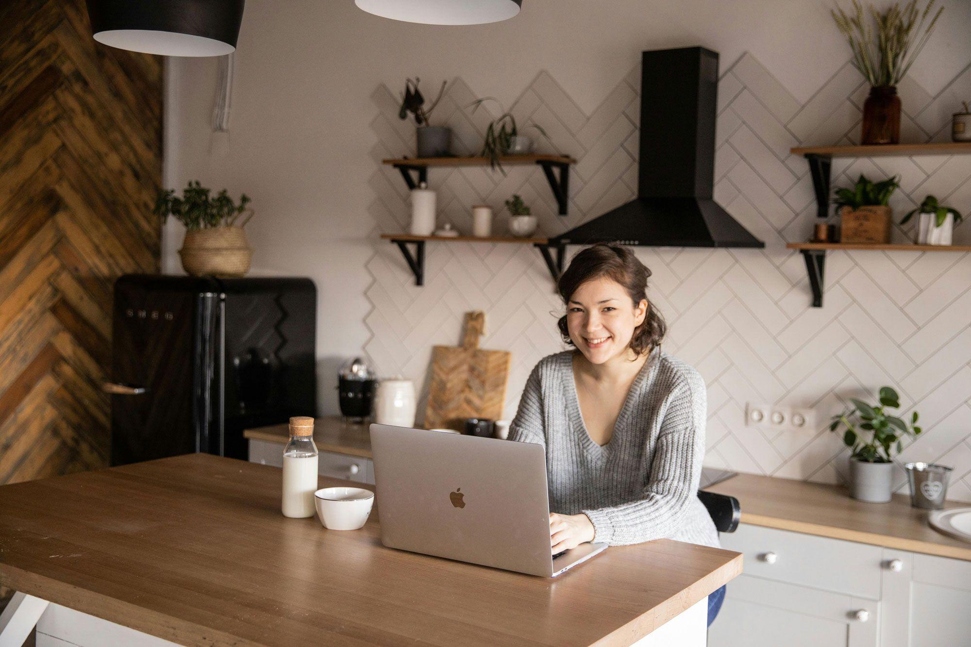 Eine Frau sitzt in einer modernen Küche an einem Holztisch mit einem Laptop, einem Glas Milch und einer Schüssel, während sie freundlich in die Kamera lächelt.