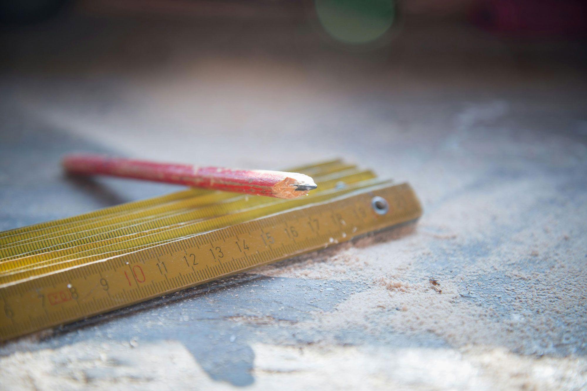 Ein roter Bleistift liegt neben einem goldfarbenen Maßstab auf einer Holzoberfläche, die mit Sägemehl bedeckt ist.