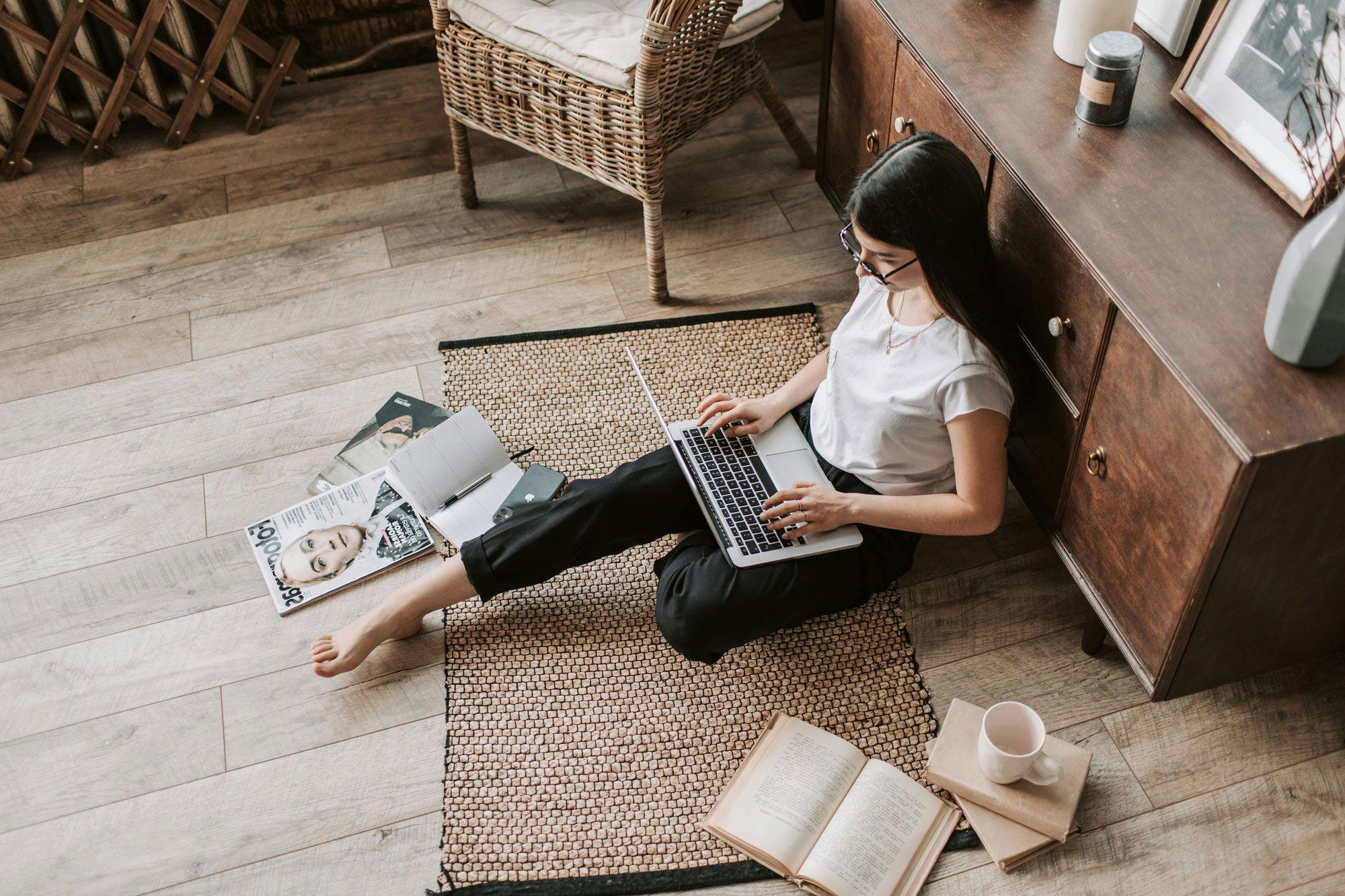 Eine Frau sitzt auf dem Boden und arbeitet an einem Laptop, umgeben von aufgeschlagenen Magazinen und einem offenen Buch, während eine Tasse Kaffee neben ihr steht.