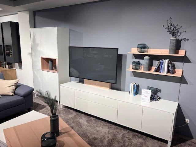 Moderne Wohnwand mit Fernseher, offenen Regalen und geschlossenen Schränken in Weiß und Holzoptik, arrangiert vor einer grauen Wand.