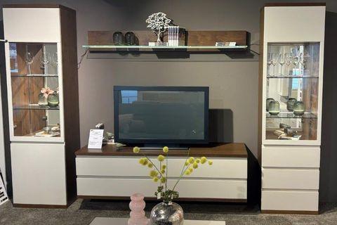 Elegante Wohnwand mit weißen Fronten und dunklen Holzakzenten. Links und rechts je eine beleuchtete Glasvitrine mit dekorativen Objekten, in der Mitte ein TV-Board mit Fernseher. Darüber ein Wandregal mit Vasen und Dekoration. Im Hintergrund eine dunkelgraue Wand.