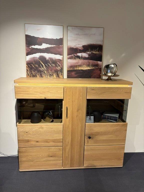 Massives Highboard aus Holz mit Glas-Elementen und vier Schubladen. Das Möbelstück ist dekoriert mit einer silbernen Vase und einer Schale auf der Oberseite. An der Wand darüber hängen zwei Kunstwerke mit Naturmotiven in warmen Farbtönen. Rechts daneben steht eine Pflanze in einem modernen, hellen Pflanzgefäß.