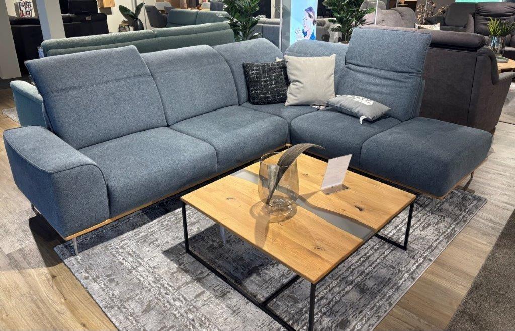 Moderne Couchgarnitur in Blau mit verstellbaren Rückenlehnen, dekoriert mit gemusterten und gestreiften Kissen. Davor ein weißer Couchtisch mit Holz- und Metallelementen, darauf eine Vase und Dekorationsobjekte. Im Hintergrund eine Vitrine und Regale aus Holz.