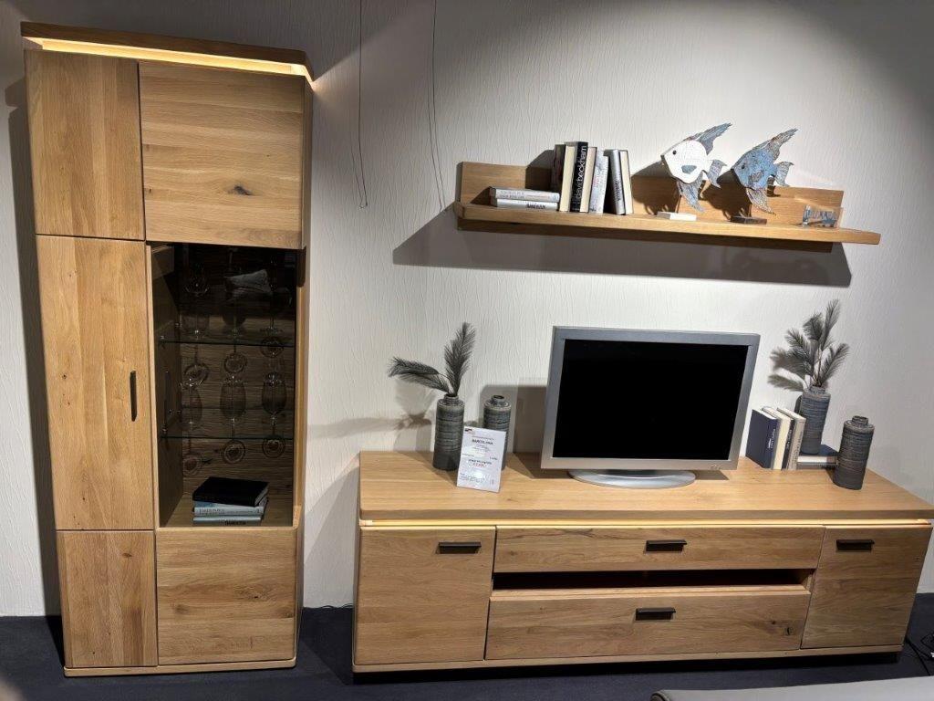 Wohnwand aus hellem Holz mit natürlicher Maserung. Links ein Schrank mit Glastür und Holzfächern, rechts ein TV-Board mit Fernseher und Schubladen. Darüber ein schlichtes Wandregal mit Büchern und dekorativen Fischen.