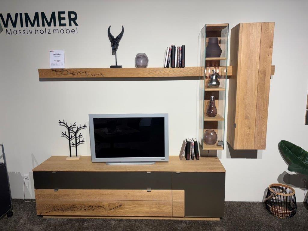 Wohnwand in modernem Design mit grauen Fronten und Holzakzenten. Links eine Glasvitrine mit dekorativen Objekten, mittig ein TV-Board mit Fernseher und Glasfach, rechts ein schwebendes Wandregal mit Büchern und Pflanzen. Dekoriert mit goldenen Kunstblättern und weiteren Accessoires.