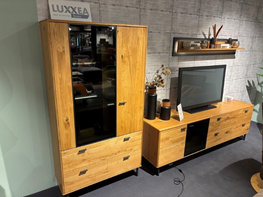 Wohnwand im rustikalen Design mit warmen Holzoberflächen. Links ein Schrank mit Glastür und Schubladen, rechts ein TV-Board mit Fernseher und dekorativer Rückwand. Darüber ein Wandregal mit Dekorationselementen.