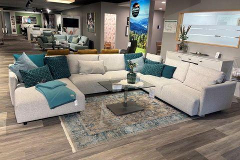 Großzügige Couchgarnitur Cielo in hellem Stoffbezug, dekoriert mit grünen und blauen Kissen, präsentiert in einem modernen Showroom.