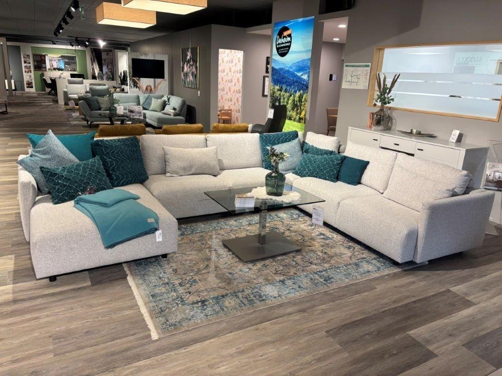 Großzügige Couchgarnitur Cielo in hellem Stoffbezug, dekoriert mit grünen und blauen Kissen, präsentiert in einem modernen Showroom.