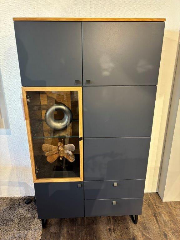 Dunkelgraue Vitrine mit Holzakzenten und einer beleuchteten Glasvitrine, die mit Gläsern dekoriert ist. Oberhalb eine große Schmetterlingsdekoration aus Holz. Steht vor einer schlichten Wand auf filigranen Metallfüßen.