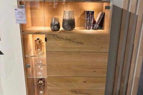 Hochwertiges Highboard aus hellem Holz mit natürlicher Maserung, links eine Glasvitrine mit beleuchteten Glasböden und Gläsern, rechts geschlossene Holzfront. Oberhalb dekoriert mit zwei Figuren und Vasen.