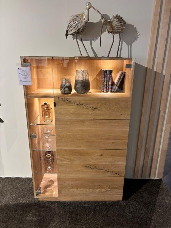 Hochwertiges Highboard aus hellem Holz mit natürlicher Maserung, links eine Glasvitrine mit beleuchteten Glasböden und Gläsern, rechts geschlossene Holzfront. Oberhalb dekoriert mit zwei Figuren und Vasen.