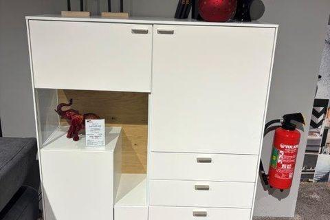 Weißes Highboard mit mehreren Schubladen und Türen, integriertem beleuchtetem Fach mit Holzrückwand, darin eine rote Elefantenfigur. Oben Dekoration mit schwarzen Vasen und einer filigranen Baumfigur. 