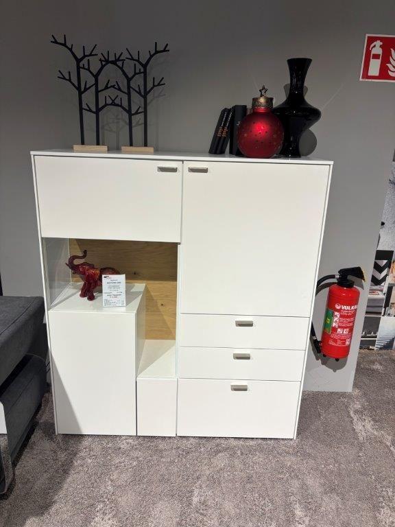 Weißes Highboard mit mehreren Schubladen und Türen, integriertem beleuchtetem Fach mit Holzrückwand, darin eine rote Elefantenfigur. Oben Dekoration mit schwarzen Vasen und einer filigranen Baumfigur. 