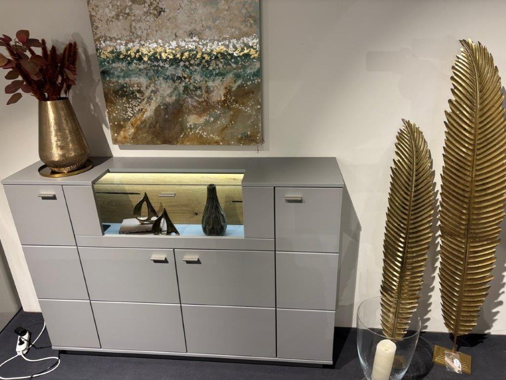 Graues Highboard mit zentralem beleuchtetem Fach, dekoriert mit Skulpturen und Vasen. Auf der Oberseite eine große goldene Vase mit roten Zweigen. Im Hintergrund ein abstraktes Gemälde mit Naturmotiven. Daneben goldene Deko-Elemente in Form von Blättern.