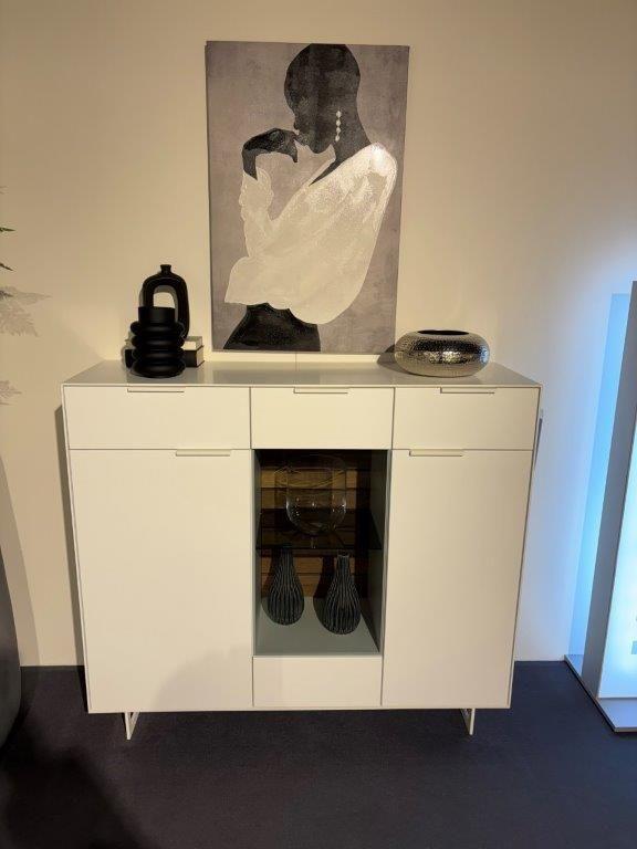 Weißes Highboard mit zentraler Glasvitrine und modernen Dekorationen, darunter Vasen und eine Schale. Daneben ein hoher grauer Pflanzentopf mit grünem Baum. Hintergrund mit heller Wand und abstraktem Kunstwerk.