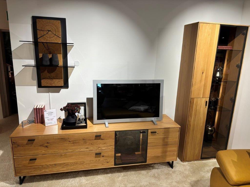 Wohnwand mit TV-Board aus hellem Holz, darauf ein Fernseher und Dekorationen. Rechts ein hohes Vitrinenschrank mit Glastüren und Beleuchtung, gefüllt mit Dekorationsgegenständen. Links ein modernes Wandregal mit schwarzem Rahmen und Holzdetails.