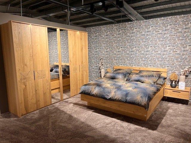 Schlafzimmer mit Kleiderschrank in Holzoptik mit Spiegeltüren, Doppelbett und Nachttischen vor einer Steinwand.