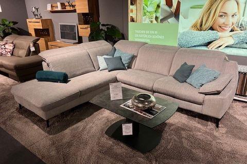 Graues Ecksofa mit blauen und grünen Kissen, kombiniert mit einem Glas-Couchtisch auf einem Teppichboden in einer modernen Wohnumgebung.