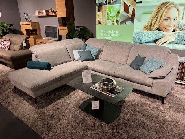 Graues Ecksofa mit blauen und grünen Kissen, kombiniert mit einem Glas-Couchtisch auf einem Teppichboden in einer modernen Wohnumgebung.