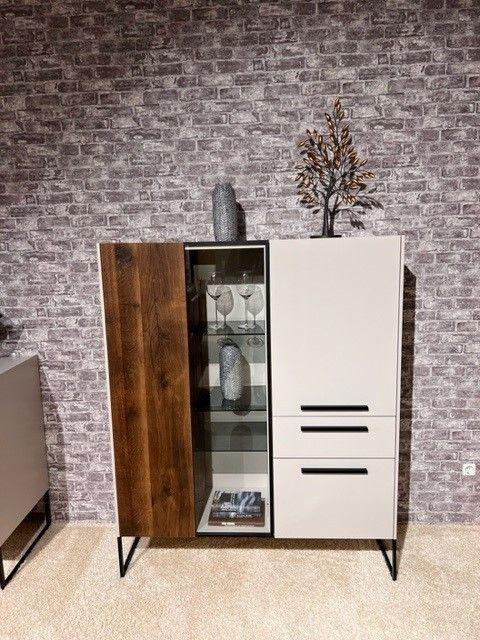 Modernes Highboard mit einer Kombination aus dunklen Holz- und weißen Fronten, einer zentralen Glasvitrine mit dekorativen Gläsern und Vasen. Steht vor einer grauen Backsteinwand und ist mit weiteren Deko-Elementen auf der Oberseite verziert.