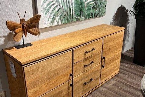 Holz-Sideboard mit schwarzen Griffen, dekoriert mit einer Holzskulptur und einer botanischen Wanddekoration, auf einem Holzboden.