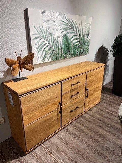 Holz-Sideboard mit schwarzen Griffen, dekoriert mit einer Holzskulptur und einer botanischen Wanddekoration, auf einem Holzboden.