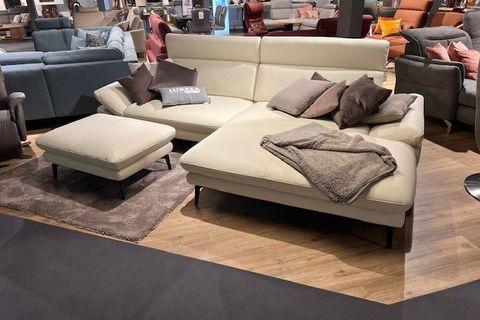 Cremefarbenes Ecksofa mit passenden Kissen und Hocker, dekoriert mit einer Decke, auf einem Holzfußboden in einer modernen Wohnumgebung.