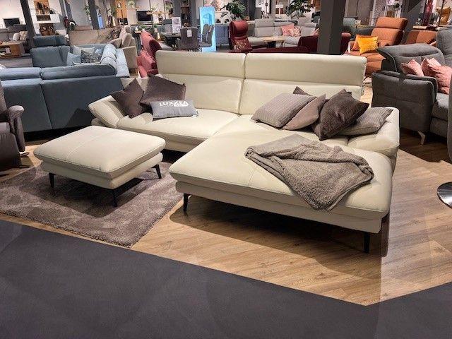 Cremefarbenes Ecksofa mit passenden Kissen und Hocker, dekoriert mit einer Decke, auf einem Holzfußboden in einer modernen Wohnumgebung.