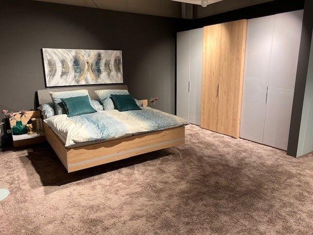 Modernes Schlafzimmer-Set mit schwebendem Bett aus hellem Holz und gepolstertem Kopfteil, dekoriert mit türkisfarbenen Kissen und passender Bettwäsche. Im Hintergrund ein großer Kleiderschrank in Weiß und Holz sowie eine Wanddekoration.