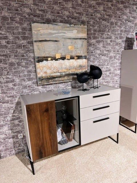 Modernes Sideboard mit weißen Fronten, dunklen Holzelementen und einer zentralen Glasvitrine, dekoriert mit Büchern und Vasen. Auf der Oberseite schwarze Skulpturen und Dekorationen. Hintergrund mit grauer Backsteinwand und abstraktem Gemälde in neutralen Farben.