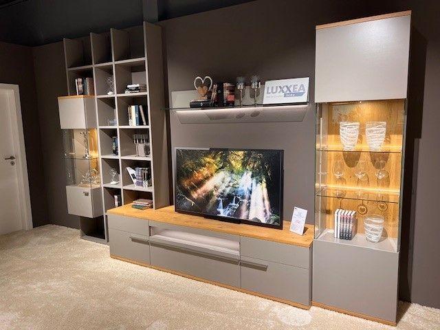 Wohnwand in modernem Design mit grauen Fronten und Holzdetails, mittig ein TV-Board mit Fernseher, links offene Regale und eine Glasvitrine, rechts eine beleuchtete Glasvitrine mit Gläsern und Dekorationen. Oben ein Wandregal mit Accessoires.