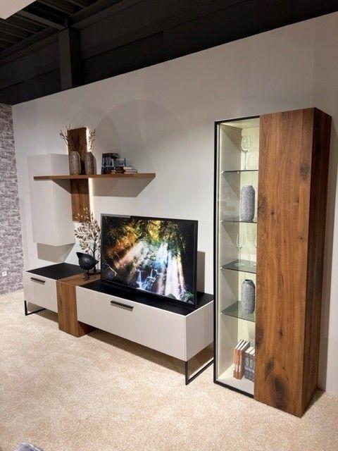 Wohnwand in modernem Design mit Holz- und weißen Elementen. Links ein TV-Board mit Schubladen und Fernseher, rechts eine hohe Glasvitrine mit Holzakzenten und dekorativen Objekten. Oben ein Wandregal mit Büchern und Vasen.