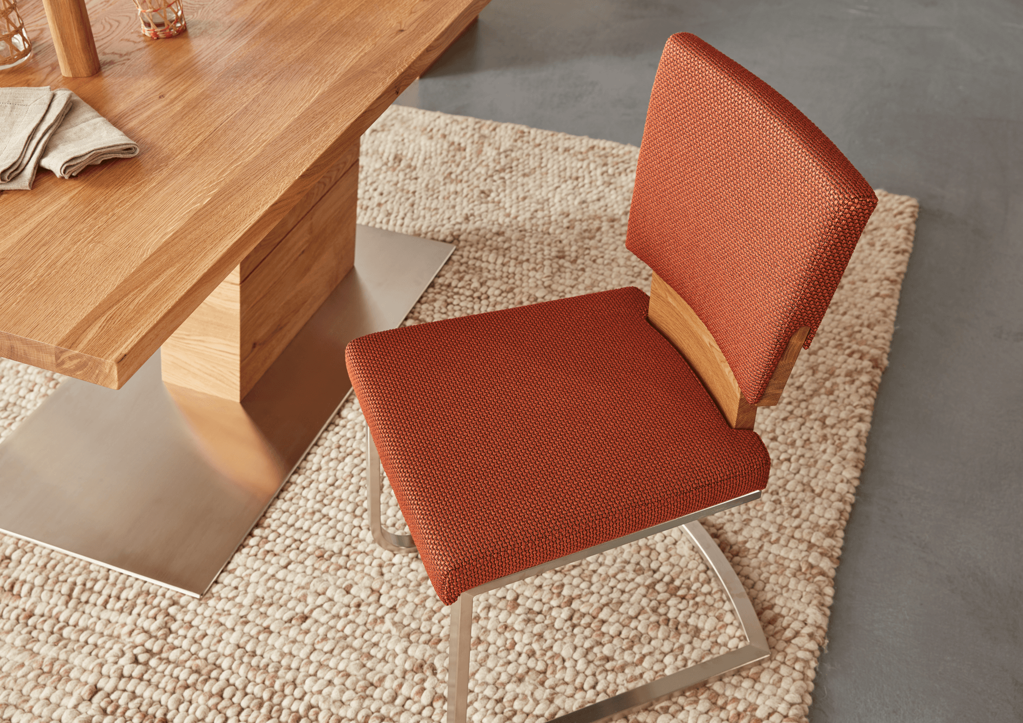 Detailaufnahme eines Essstuhls mit rotem Polsterbezug und Holzrückenlehne, kombiniert mit einem massiven Holztisch auf Metallfuß; arrangiert auf einem strukturierten Teppich.