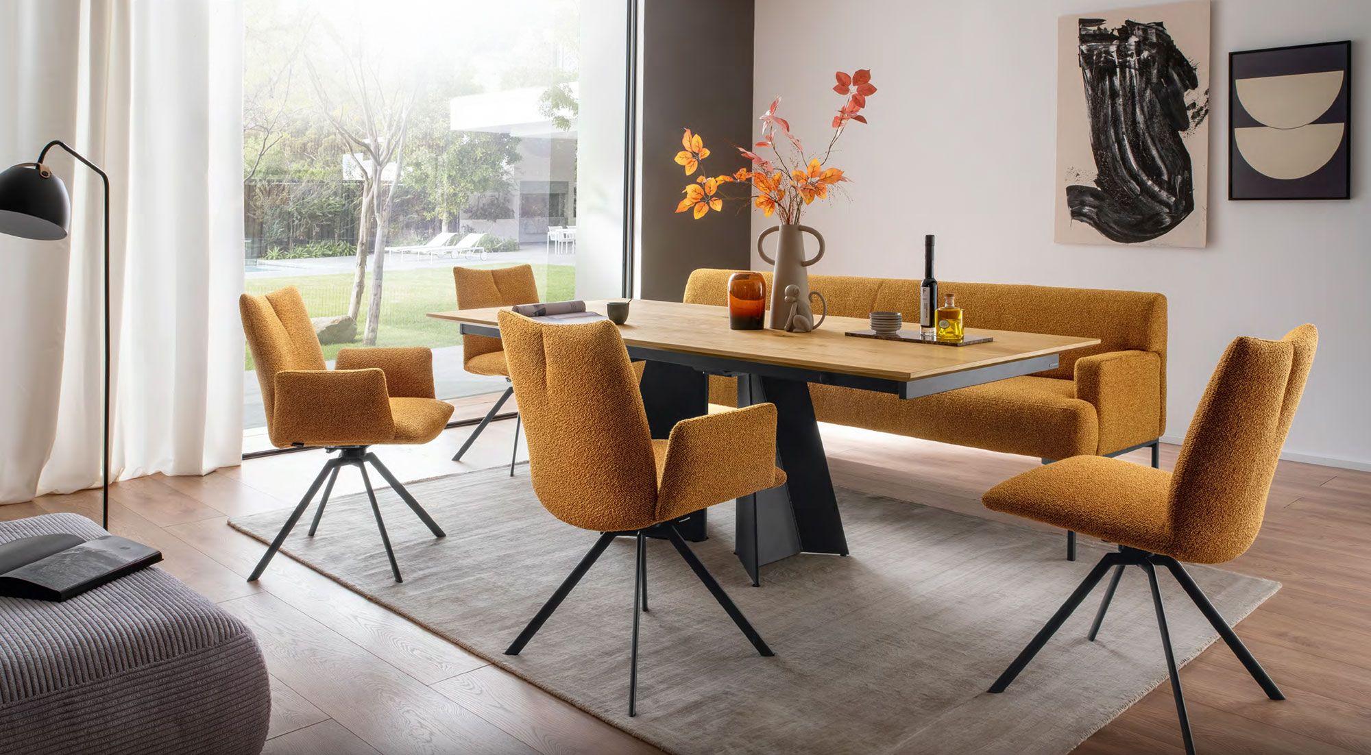 Modernes Esszimmer-Set mit einem Holztisch und orangefarbenen Polsterstühlen sowie einer passenden Bank. Dekoratives Highlight sind herbstliche Blumen in einer Vase auf dem Tisch.