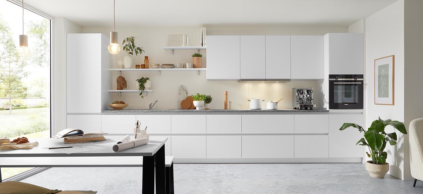 Helle, minimalistische Küche in Weiß mit grauer Arbeitsplatte, offenen Regalen und stilvollen Dekorationen; große Fenster sorgen für viel Tageslicht und eine freundliche Atmosphäre.