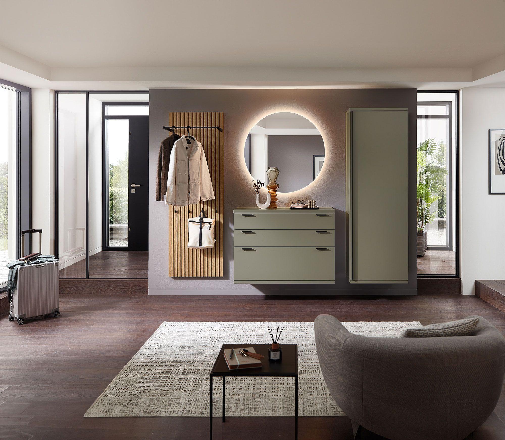 Moderner Eingangsbereich mit hängenden Möbeln in Salbeigrün, einer vertikalen Garderobenleiste aus Holzlamellen und einem großen, runden Spiegel mit integrierter Beleuchtung. Dunkler Holzboden und ein heller Teppich ergänzen das elegante Design.