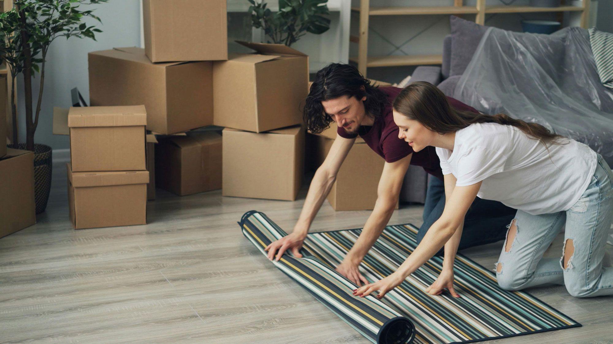 Ein Paar, das gemeinsam einen gestreiften Teppich auf dem Boden eines neu eingerichteten Raumes ausrollt, umgeben von Umzugskartons. Ein Paar, das gemeinsam einen gestreiften Teppich auf dem Boden eines neu eingerichteten Raumes ausrollt, umgeben von Umzugskartons.