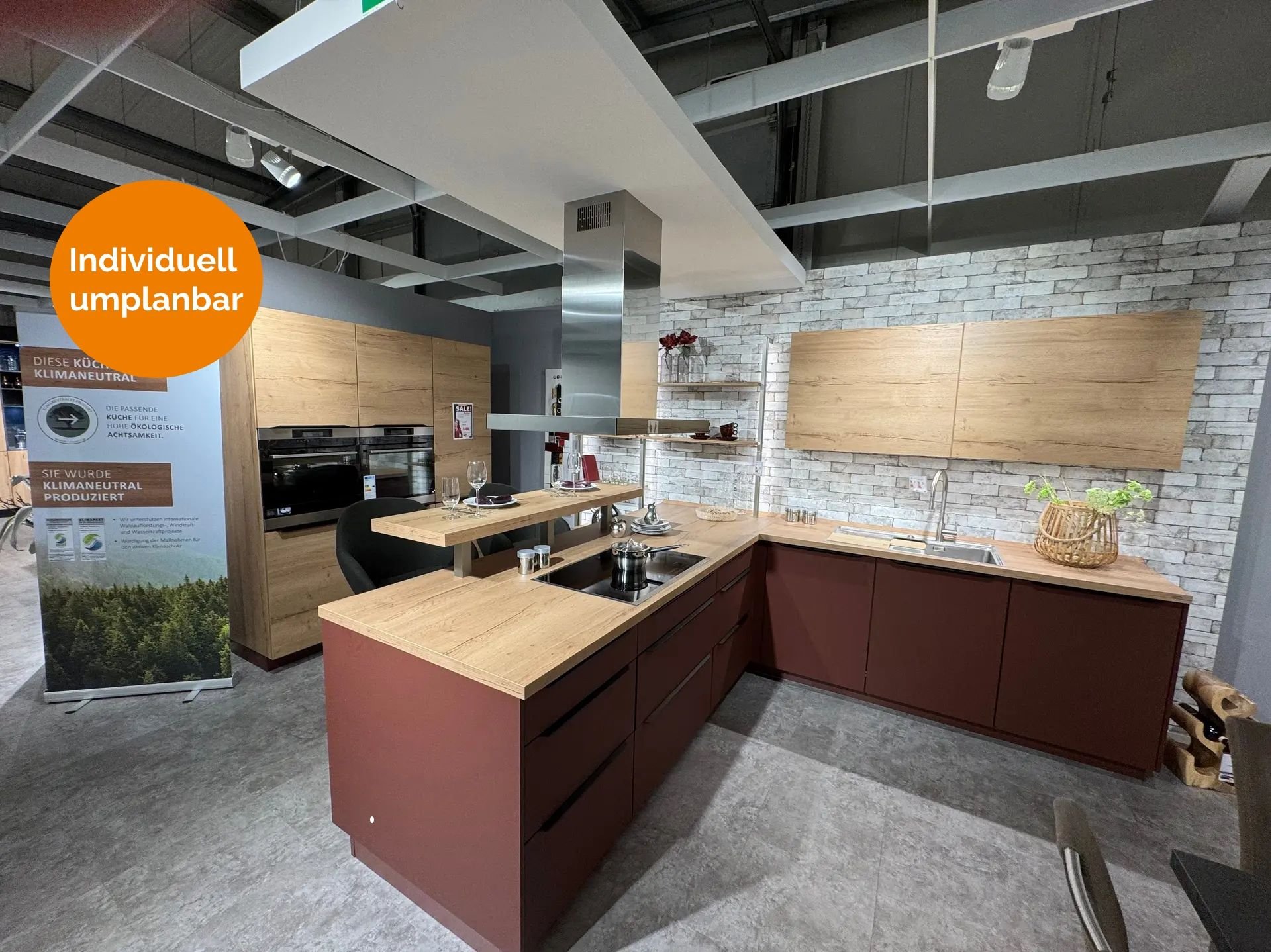 Klimaneutrale Küche in Terrakottarot und Eiche-Optik mit Kochinsel, integriertem Essplatz, Edelstahl-Inselhaube, Steinwand-Verblender und Hochschrank mit Einbaugeräten.