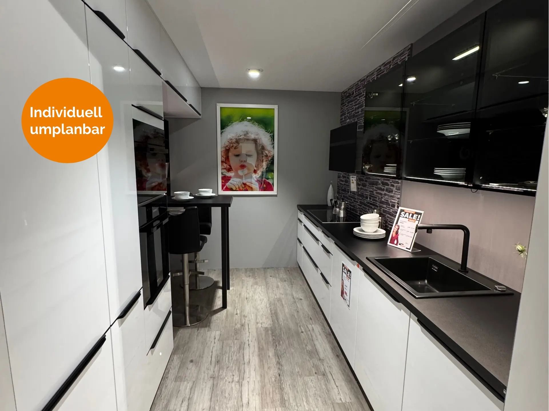 Küche in Weiß Hochglanz mit schwarzen Griffleisten, dunkler Arbeitsplatte, schwarzen Glas-Hängeschränken mit Innenbeleuchtung, Steinoptik-Nischenrückwand, schwarzer Armatur und kleinem Bartisch mit Hockern