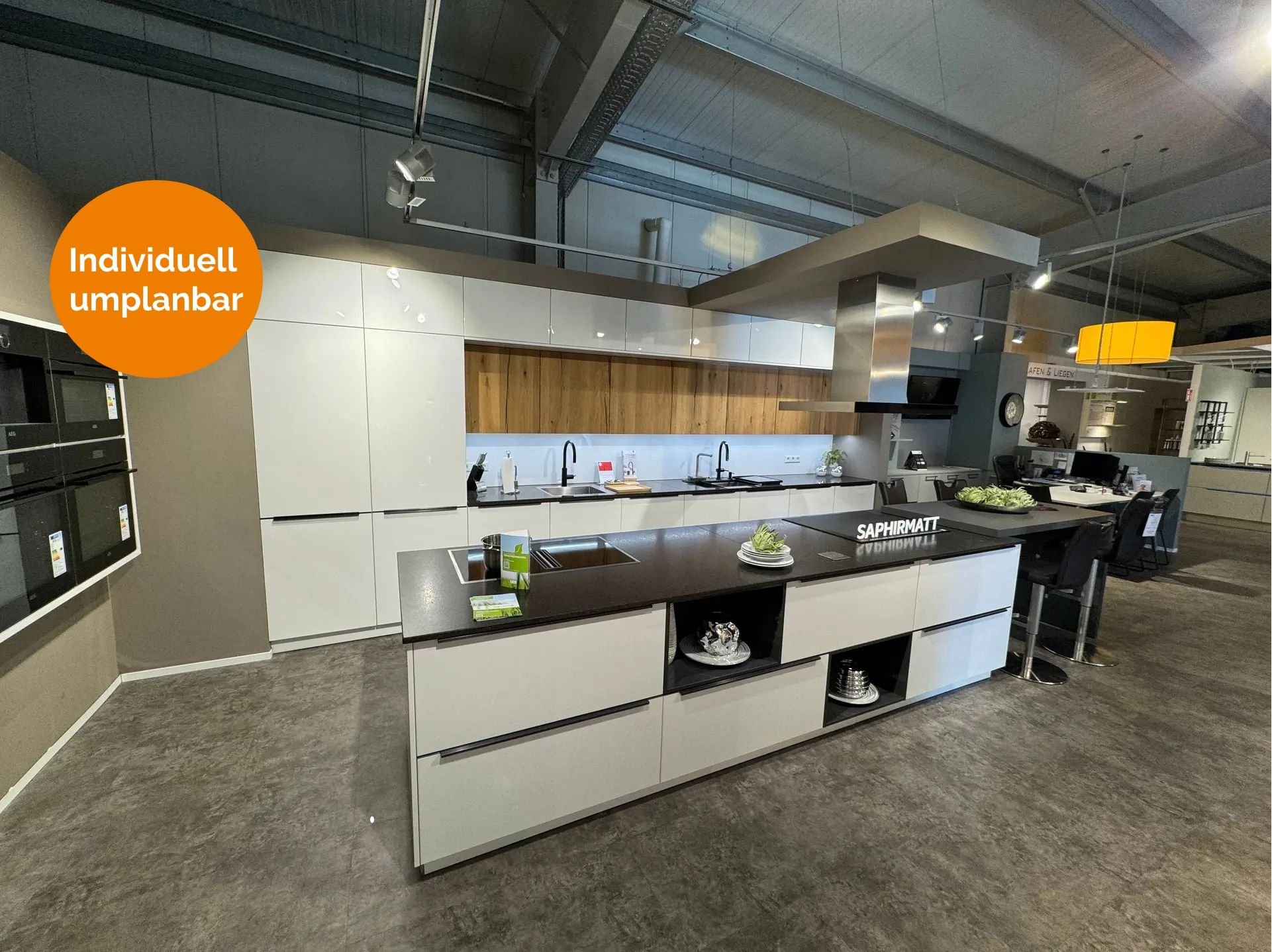 Große Küche in Saphirmatt Weiß mit dunkler Arbeitsplatte, Kochinsel mit Kochfeld und offenen Designfächern, Holz-Nischenrückwand in Eiche, grifflosen Fronten, Deckenlifthaube und integriertem Essplatz mit Barhockern.