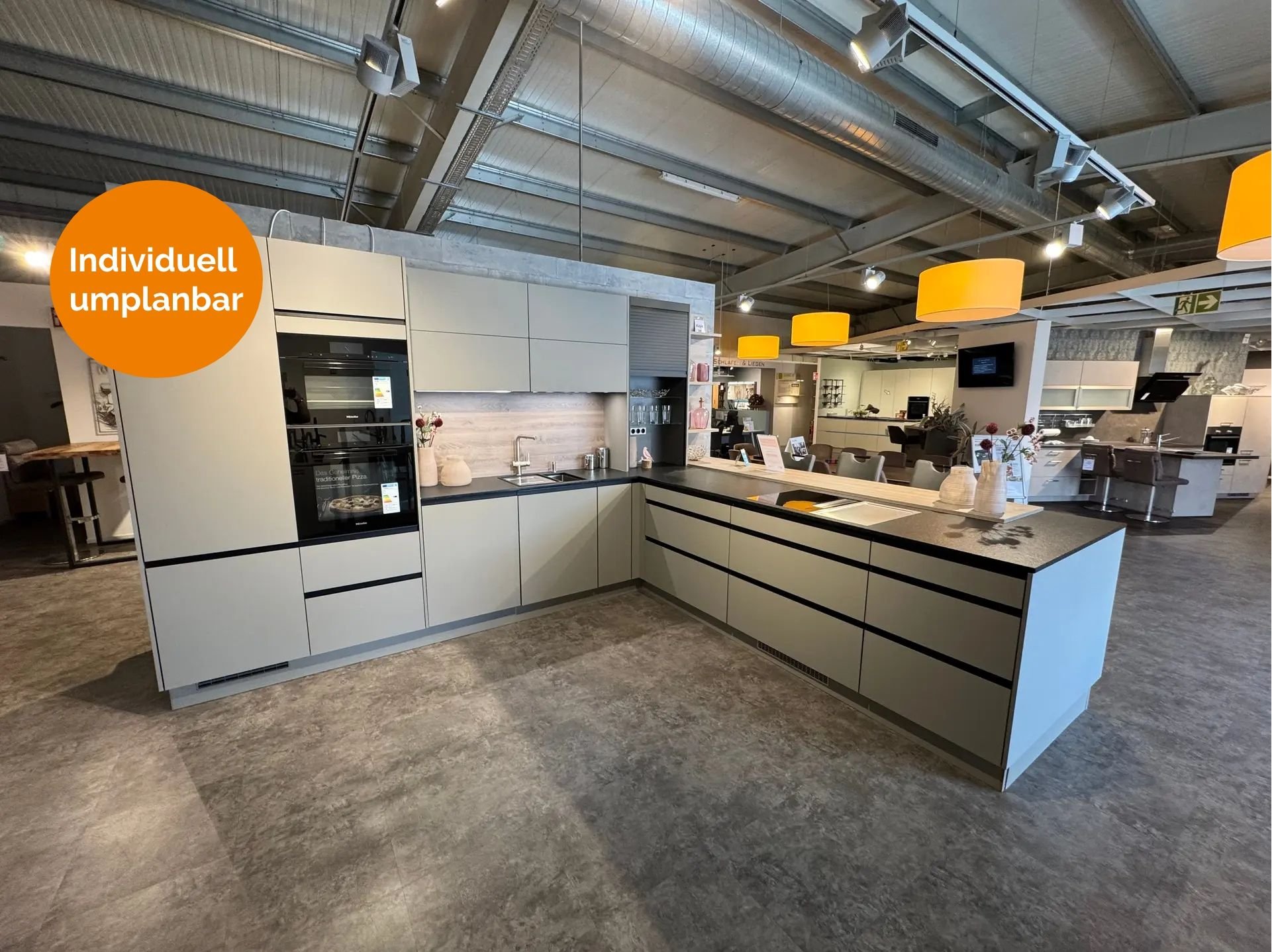 Großzügige U-Küche in mattem Graubeige mit schwarzen Griffmulden, dunkler Arbeitsplatte, Holz-Nischenrückwand, Hochschrank mit Miele-Einbaugeräten und Halbinsel mit Kochfeld.