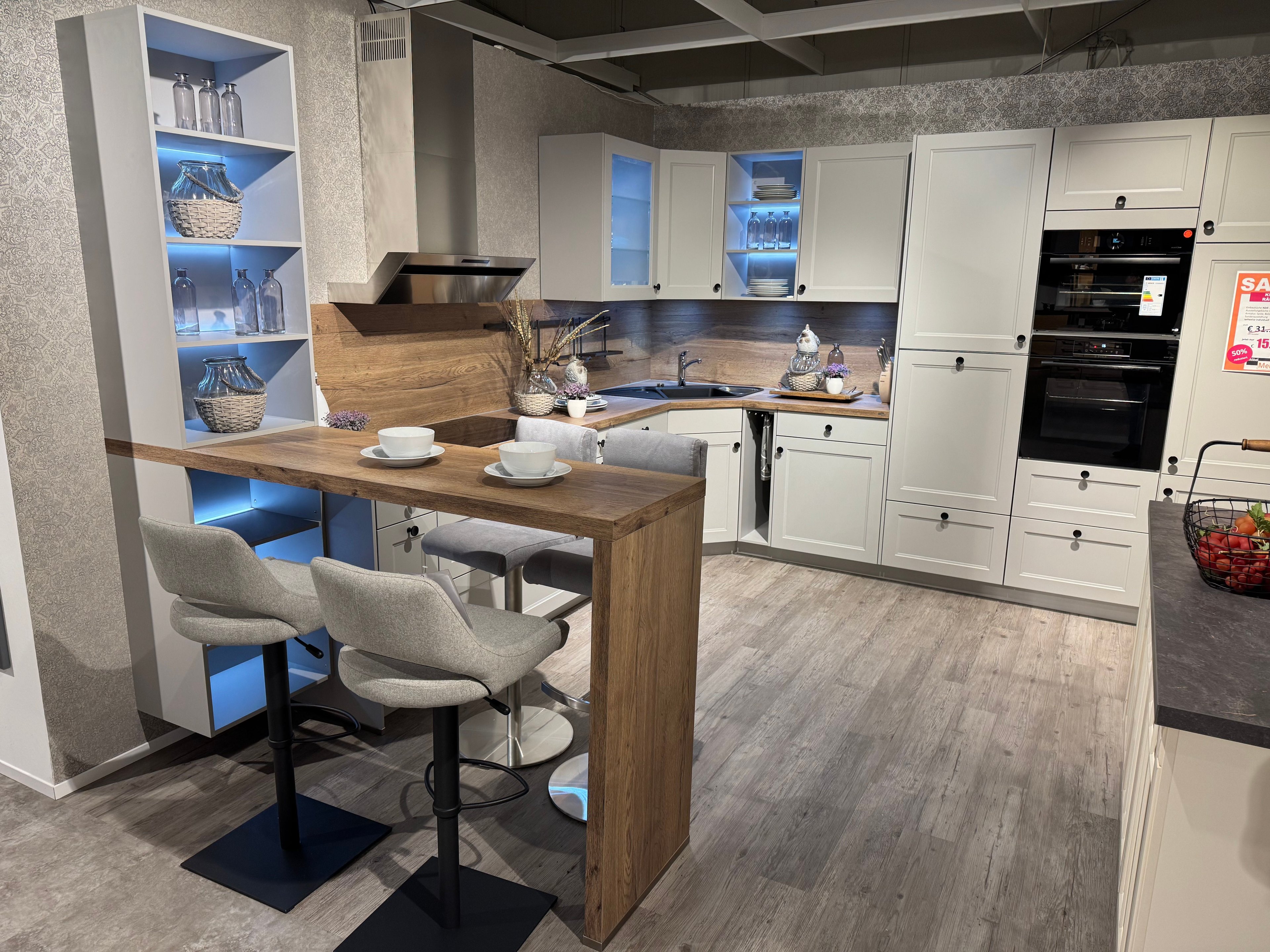 Weiße Landhausküche mit Rahmenfronten, Holzarbeitsplatte und Nischenrückwand in Eiche, Bartresen mit Polsterhockern, beleuchtetem Designregal mit blauer LED-Beleuchtung und Hochschrank mit Einbaugeräten.